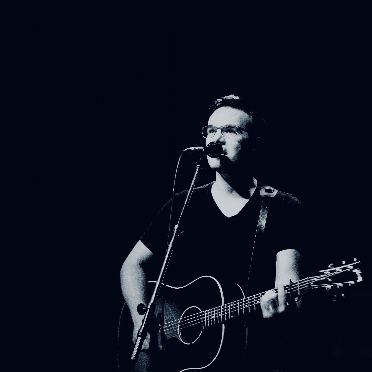 Schwarzweiß-Porträt: Simon Schütt singt ins Mikrofon und spielt Gitarre vor dunklem Hintergrund