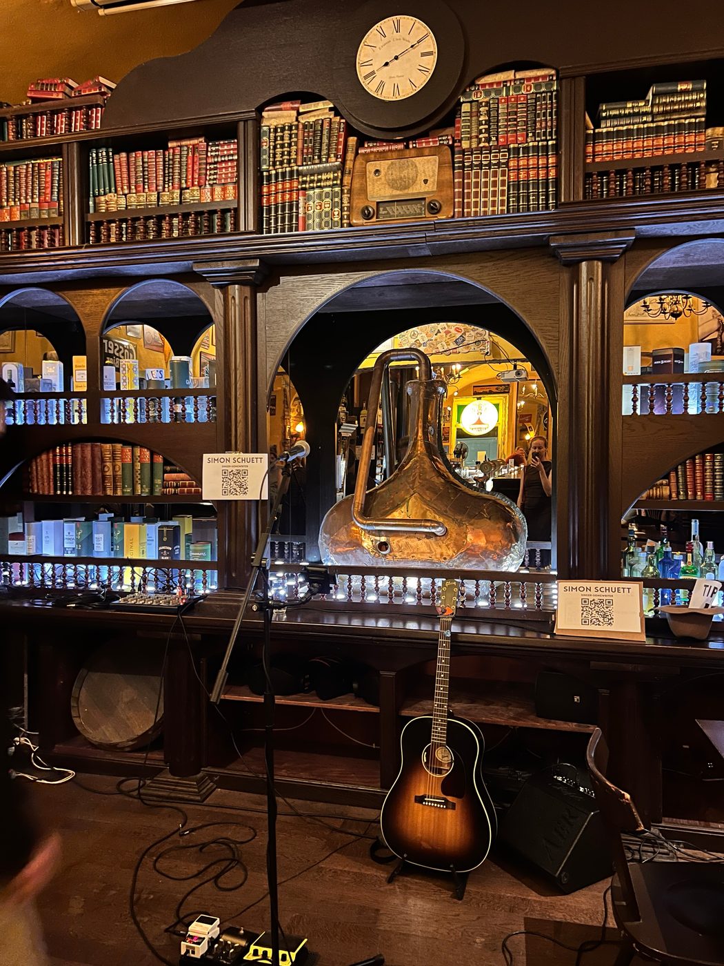 Simons Setup in einem Pub: Akustikgitarre auf dem Ständer, Mikrofonständer, kleines Mischpult, Pedale und Monitor vor einer Regalwand